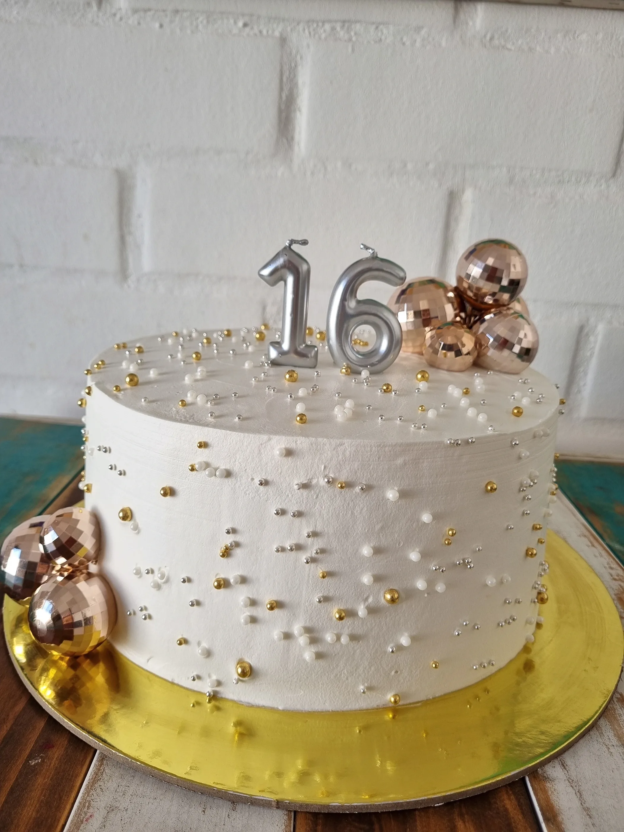 Torta redonda blanca con perlas doradas, número 16 plateado y esferas metálicas sobre base dorada.