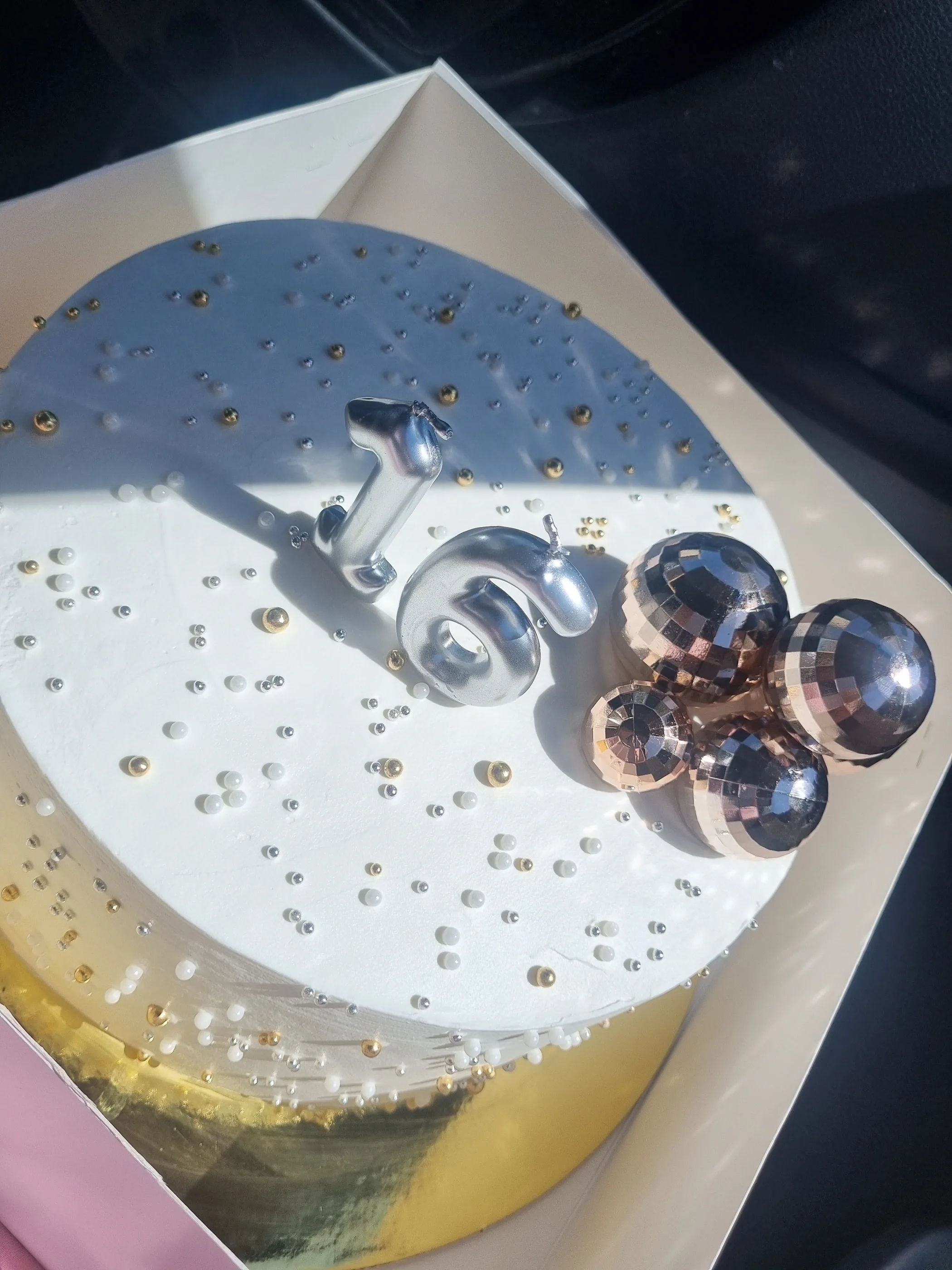 Vista superior de torta blanca con número 16, perlas y esferas metálicas sobre base dorada.