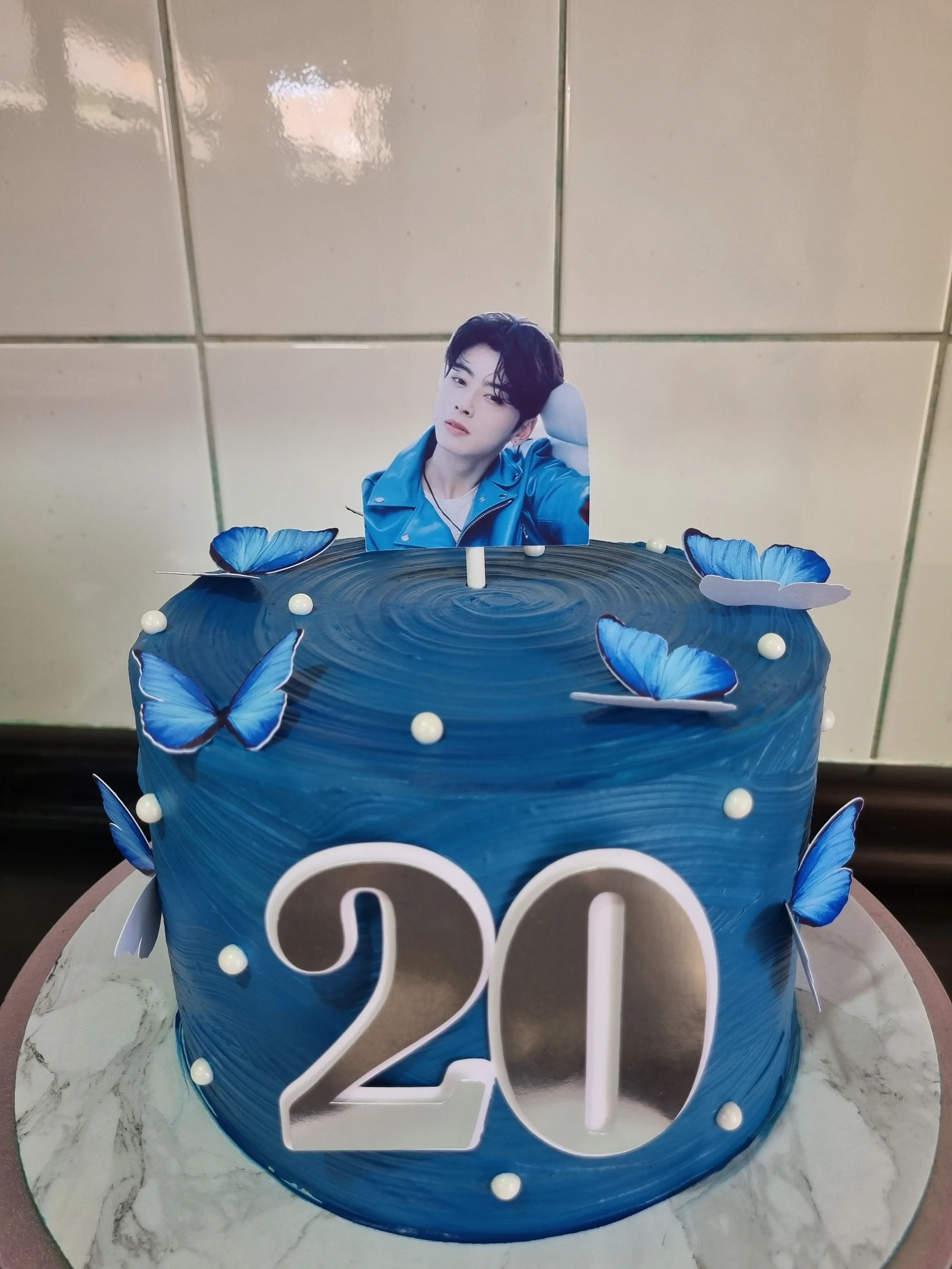 Torta azul con mariposas, perlas blancas, número 20 y topper fotográfico de un cantante.