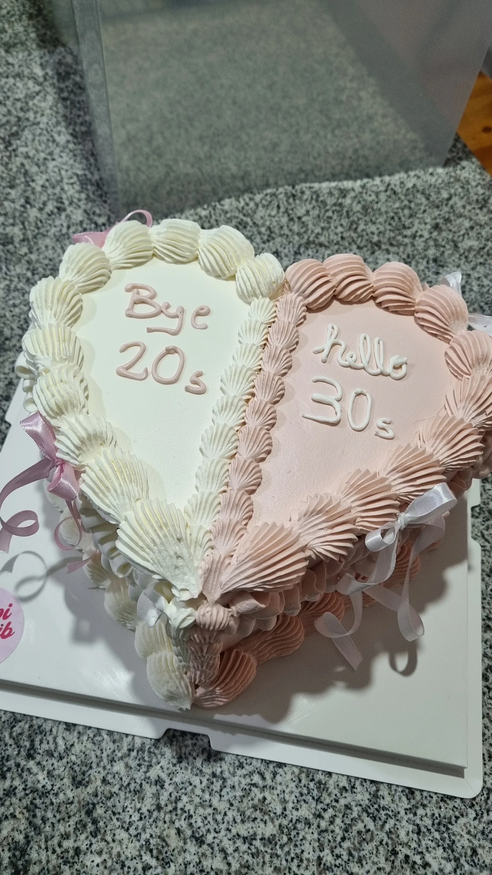 Torta en forma de corazón, mitad marfil y mitad rosa, con bordes de crema e inscripciones Bye 20s y Hello 30s.