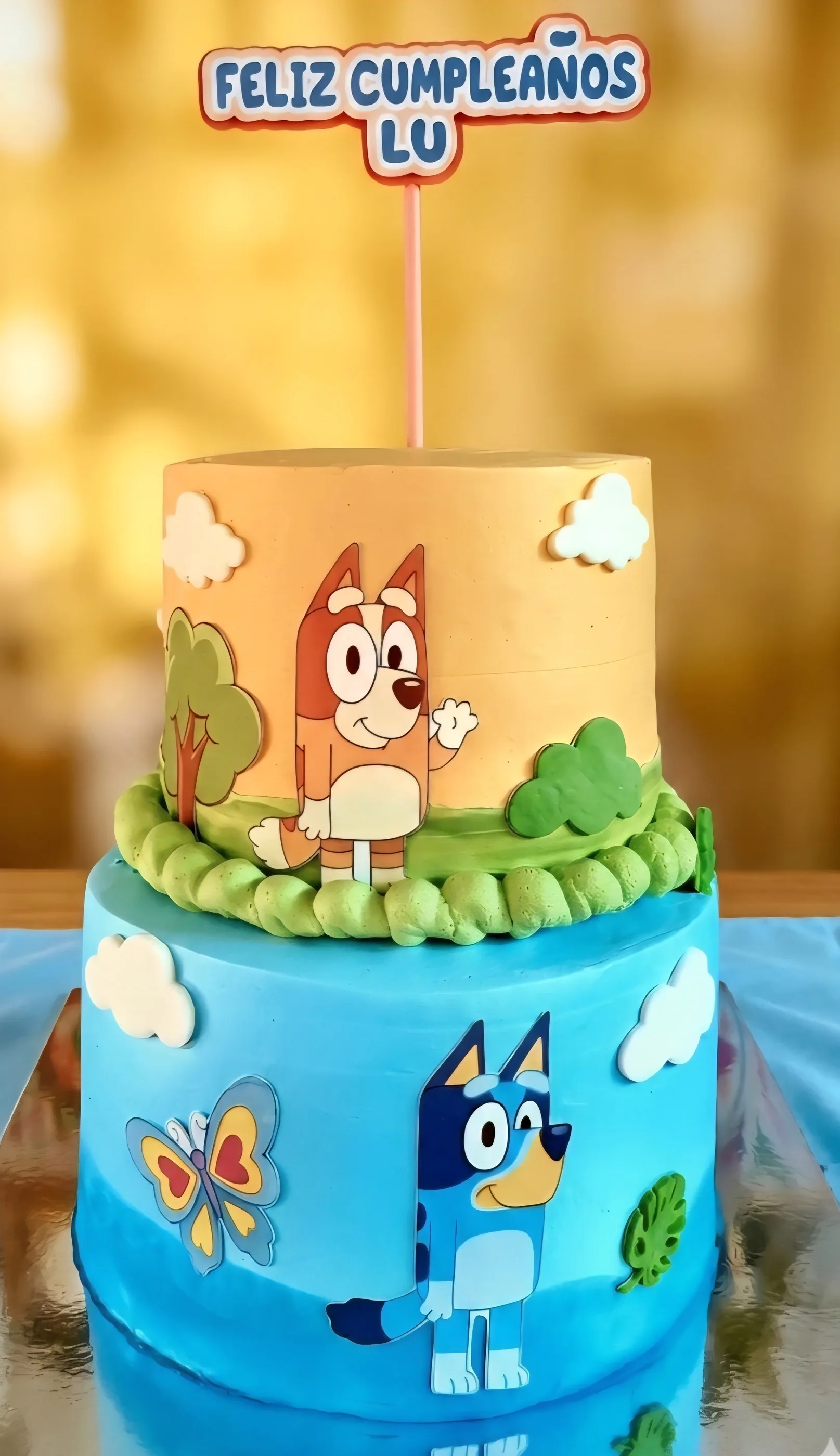 Torta de dos pisos naranja y celeste con Bluey y Bingo, nubes, flores y letrero de cumpleaños.