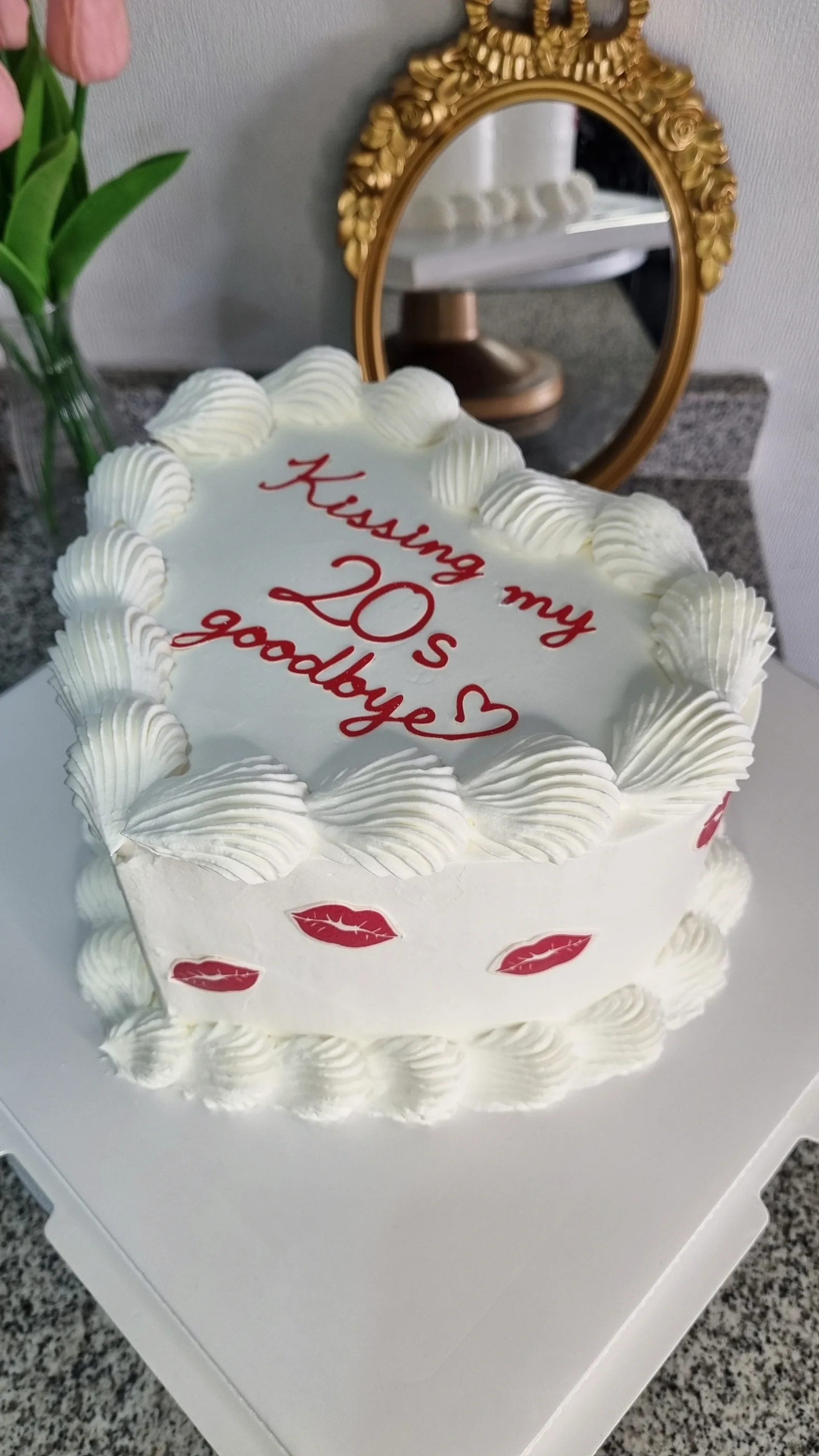 Torta blanca con bordes de crema e inscripción roja Kissing my 20s goodbye y pequeños besos rojos.