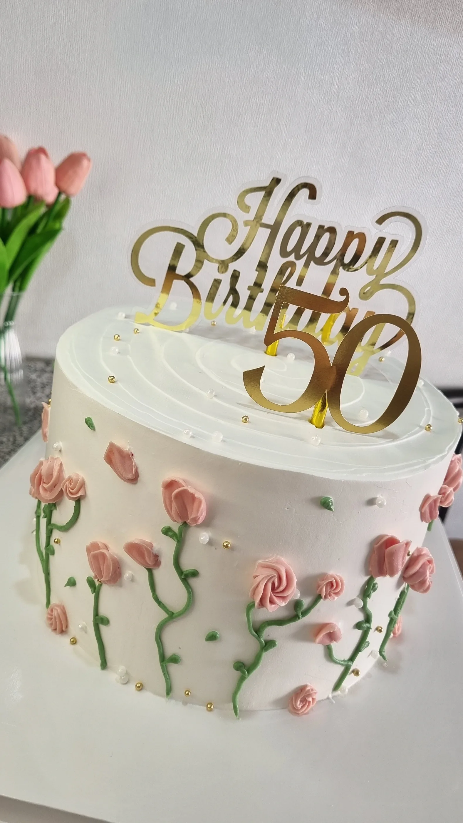 Torta blanca con rosas pequeñas en relieve, tallos verdes y letrero dorado Happy Birthday 50.