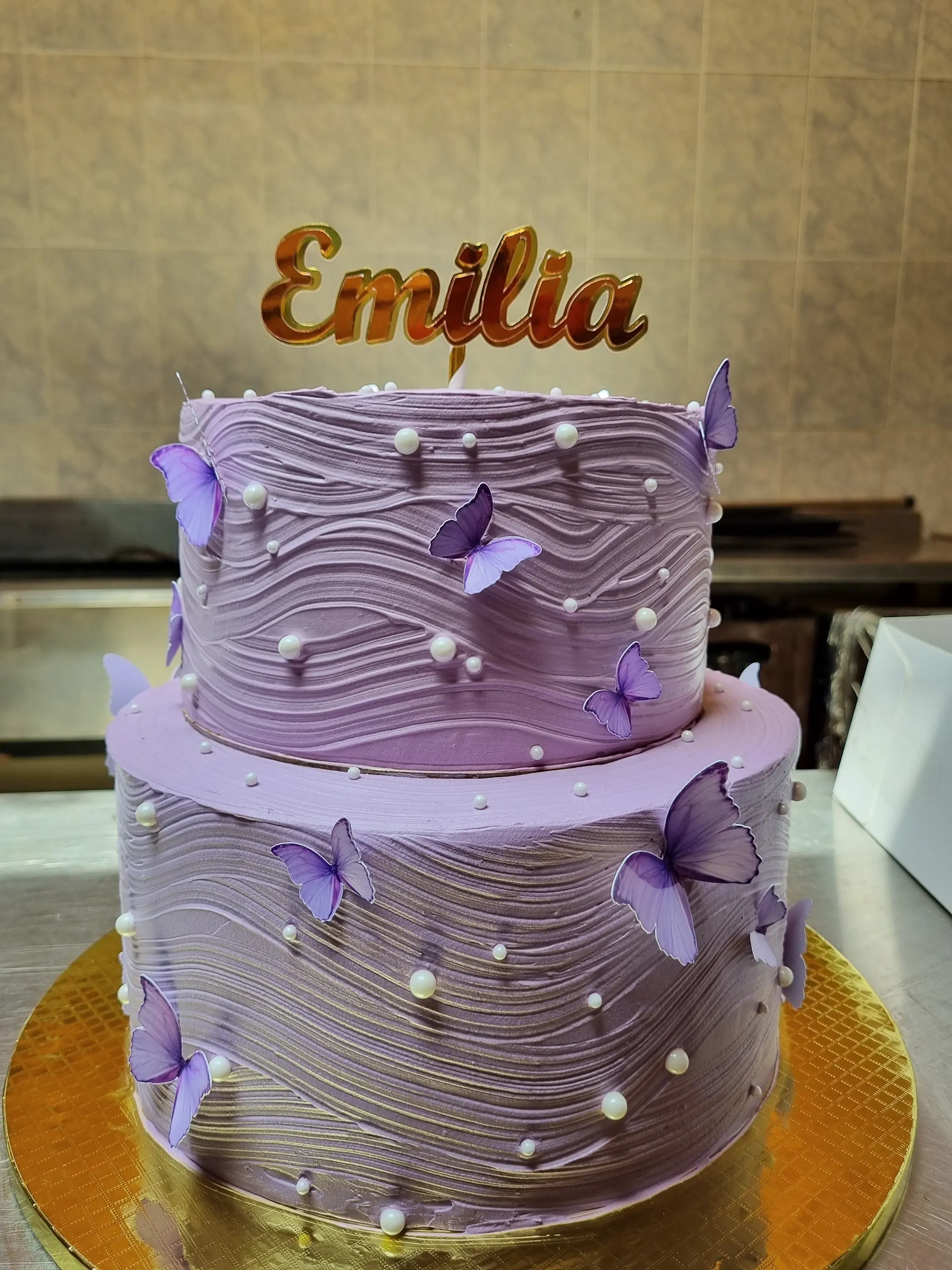 Torta lila de dos pisos con ondas, perlas blancas, mariposas violetas y nombre Emilia.
