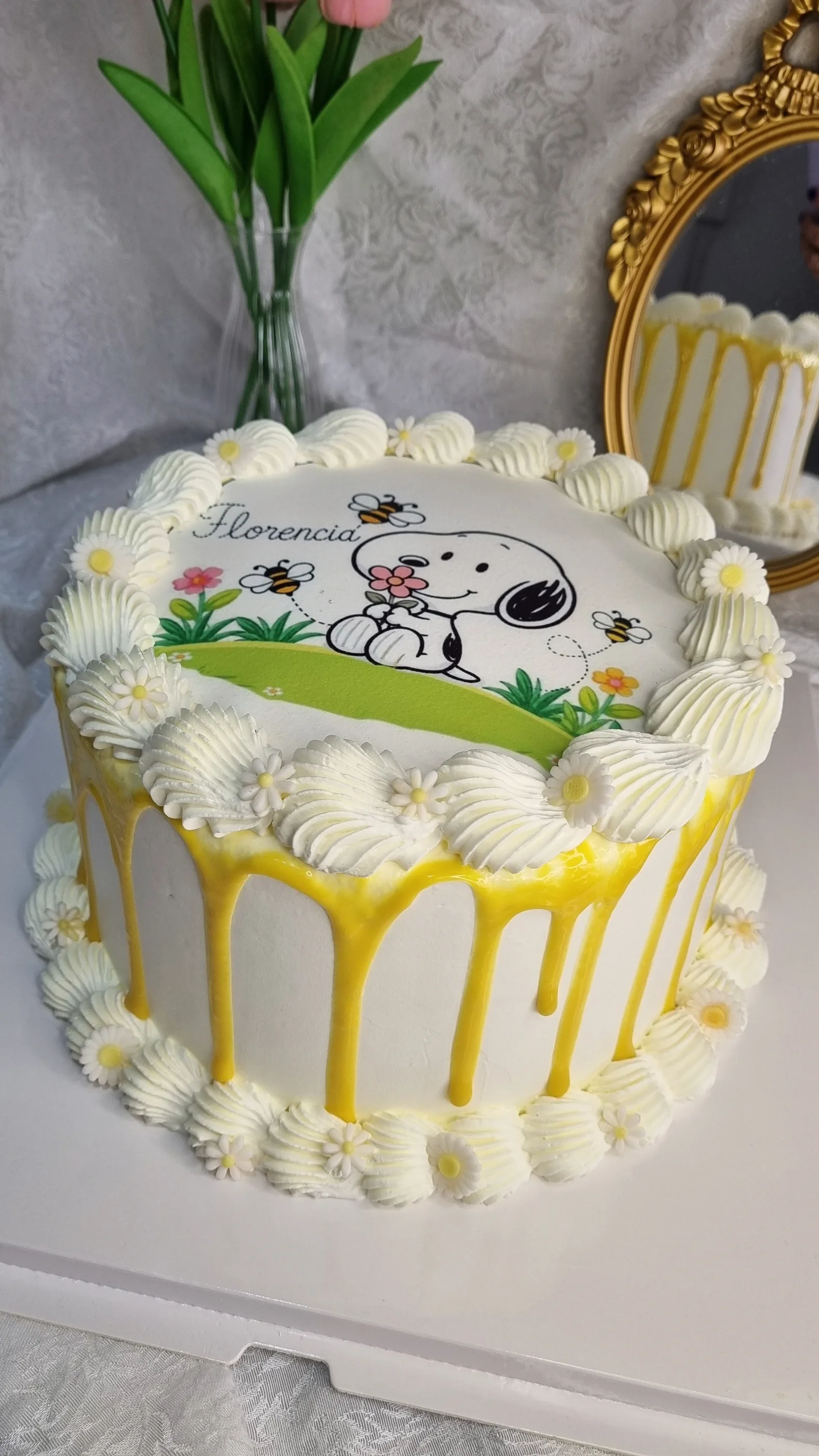 Torta blanca con ilustración de Snoopy, goteado amarillo, bordes de crema y margaritas.