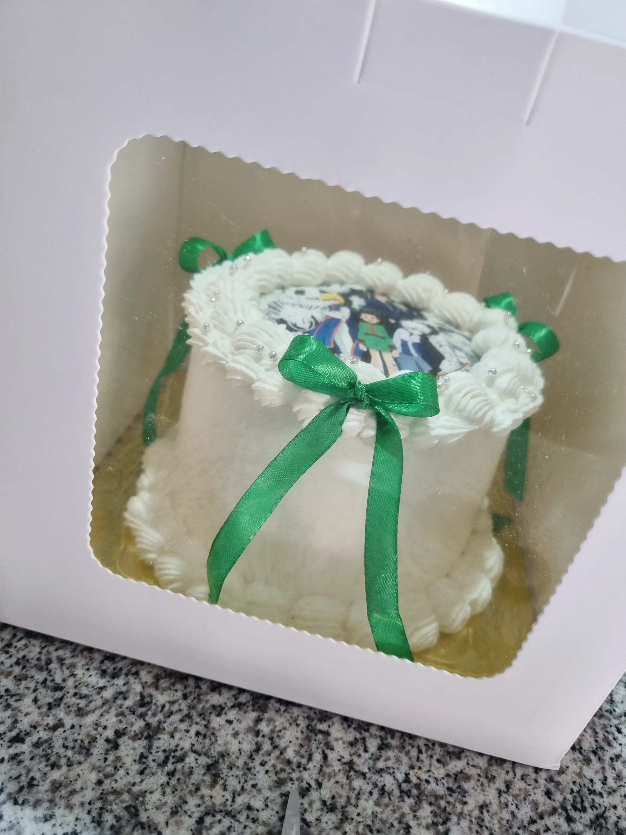 Torta blanca con lazo verde y decoración impresa, visible a través de una caja con ventana.