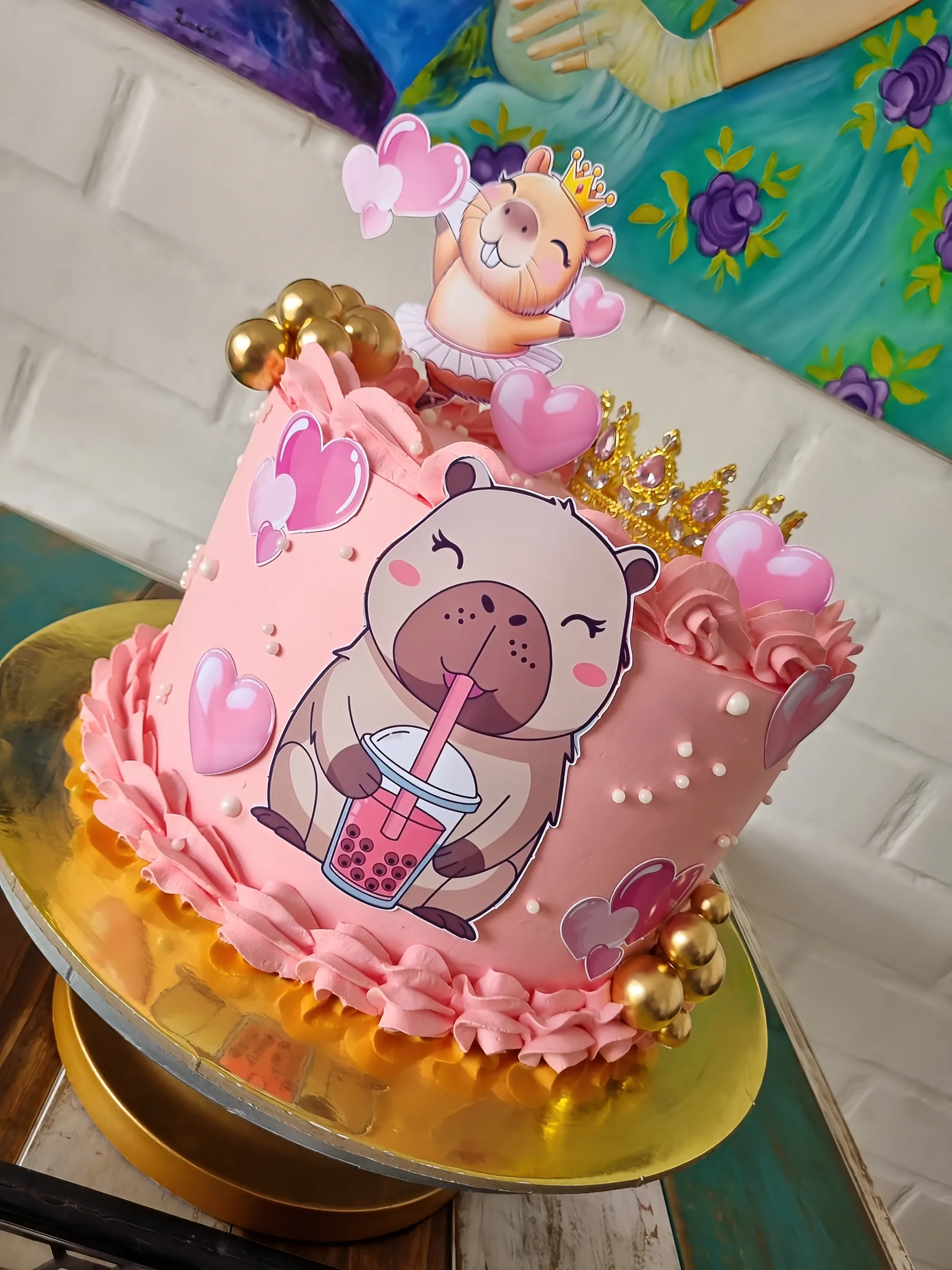 Torta rosa con capibara sosteniendo una bebida, corazones, perlas y esferas doradas.
