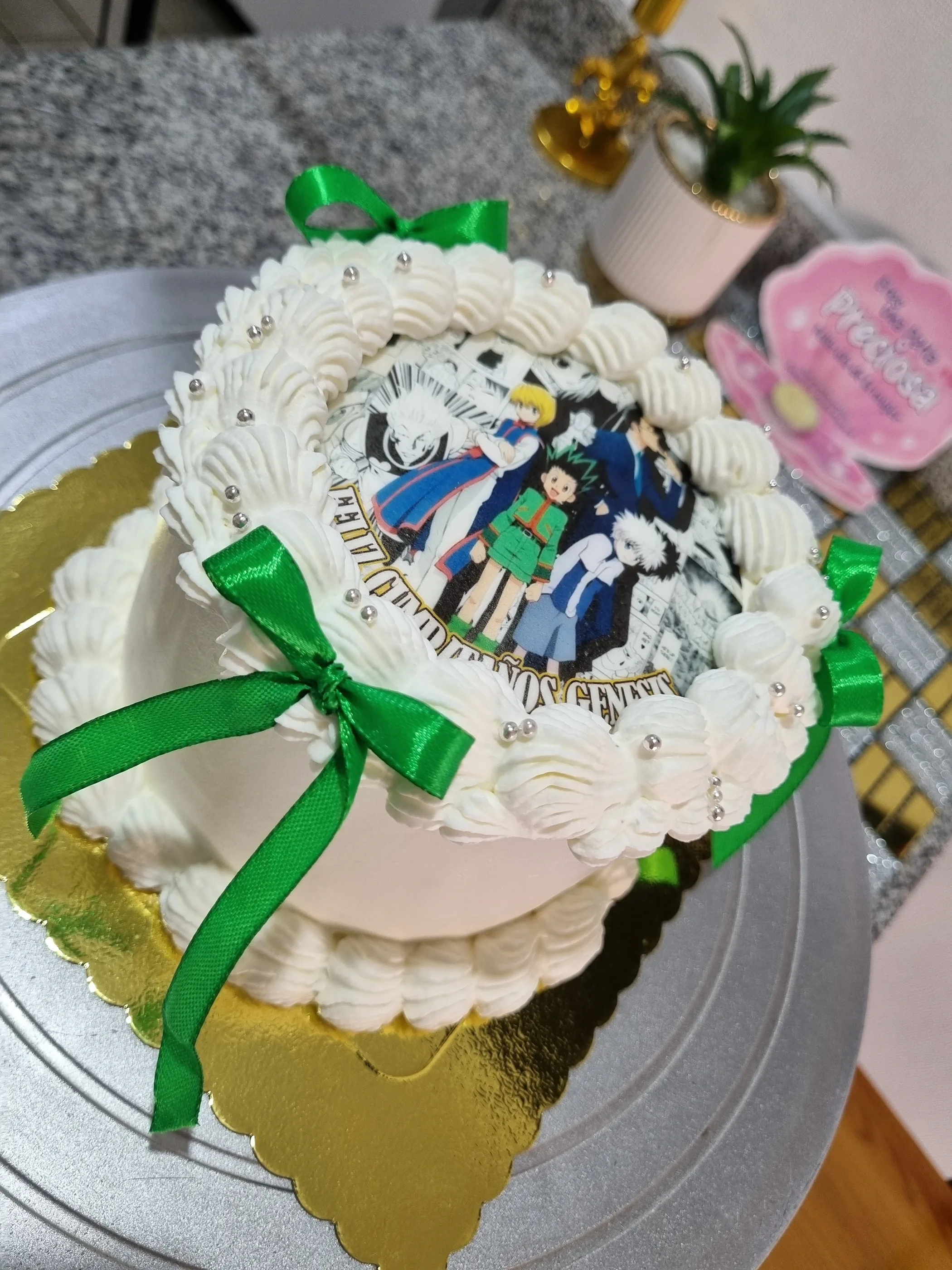 Torta blanca con imagen impresa de personajes de anime y lazos verdes sobre base dorada.