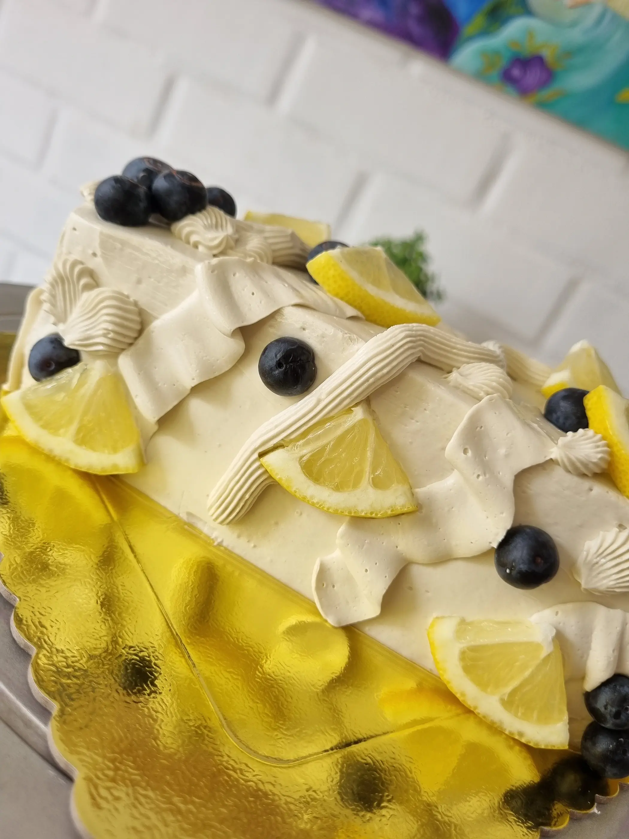 Torta decorada con pliegues de crema, rodajas de limón y arándanos sobre base dorada.