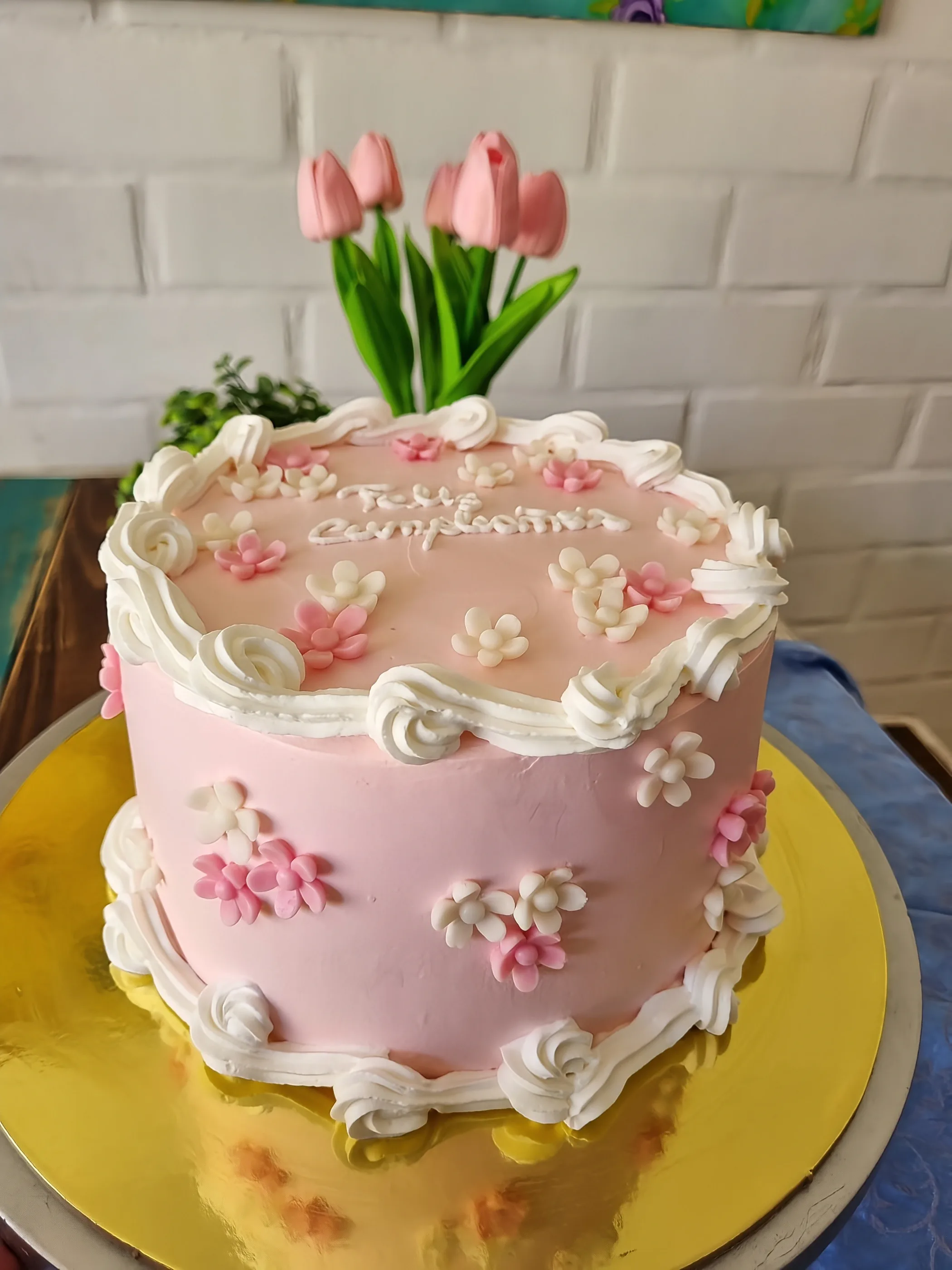 Torta rosa con pequeñas flores blancas y rosadas, bordes de crema y mensaje de cumpleaños.