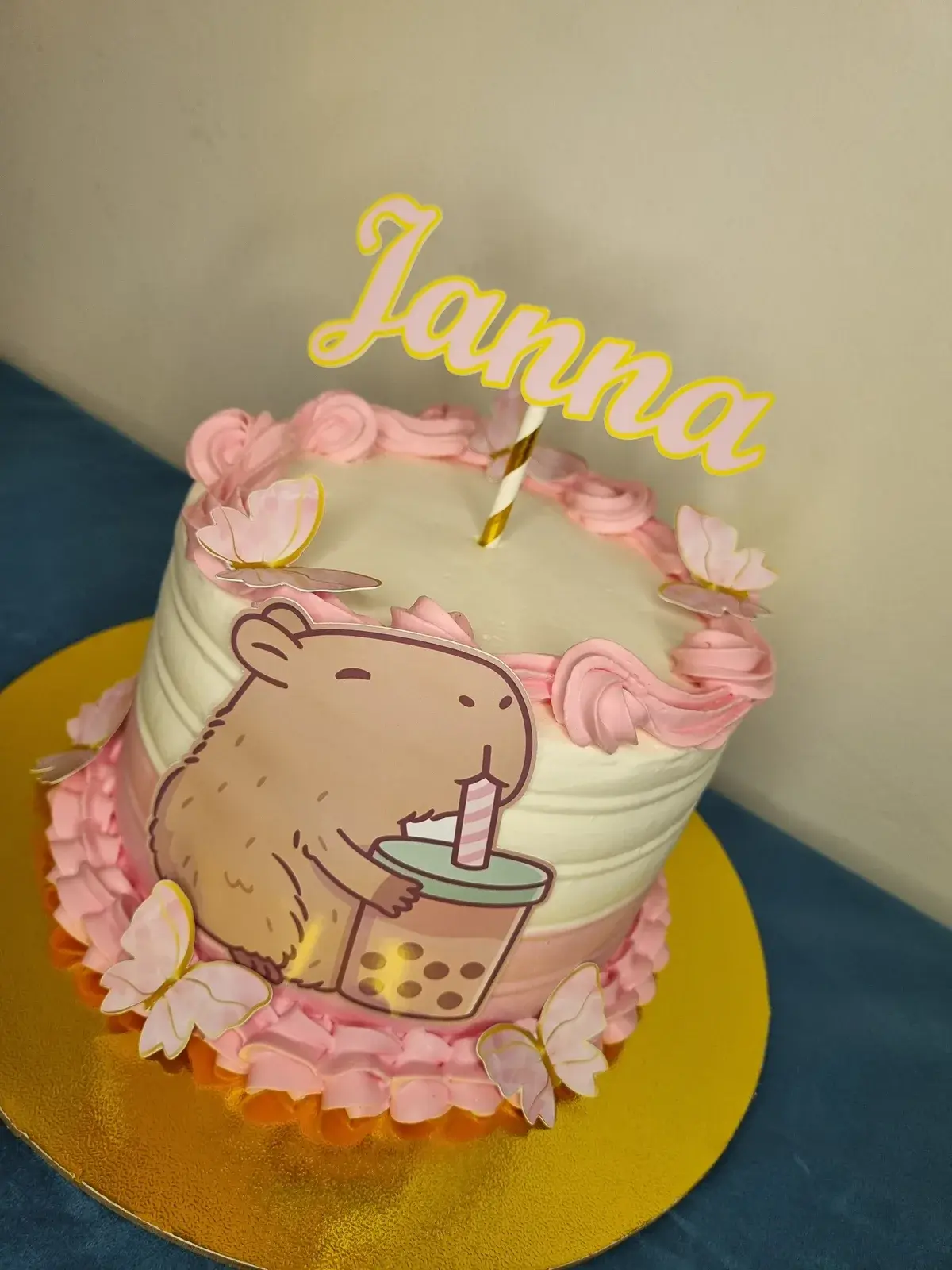Torta blanca con bordes rosados, topper de capibara, mariposas y nombre Janna en dorado.