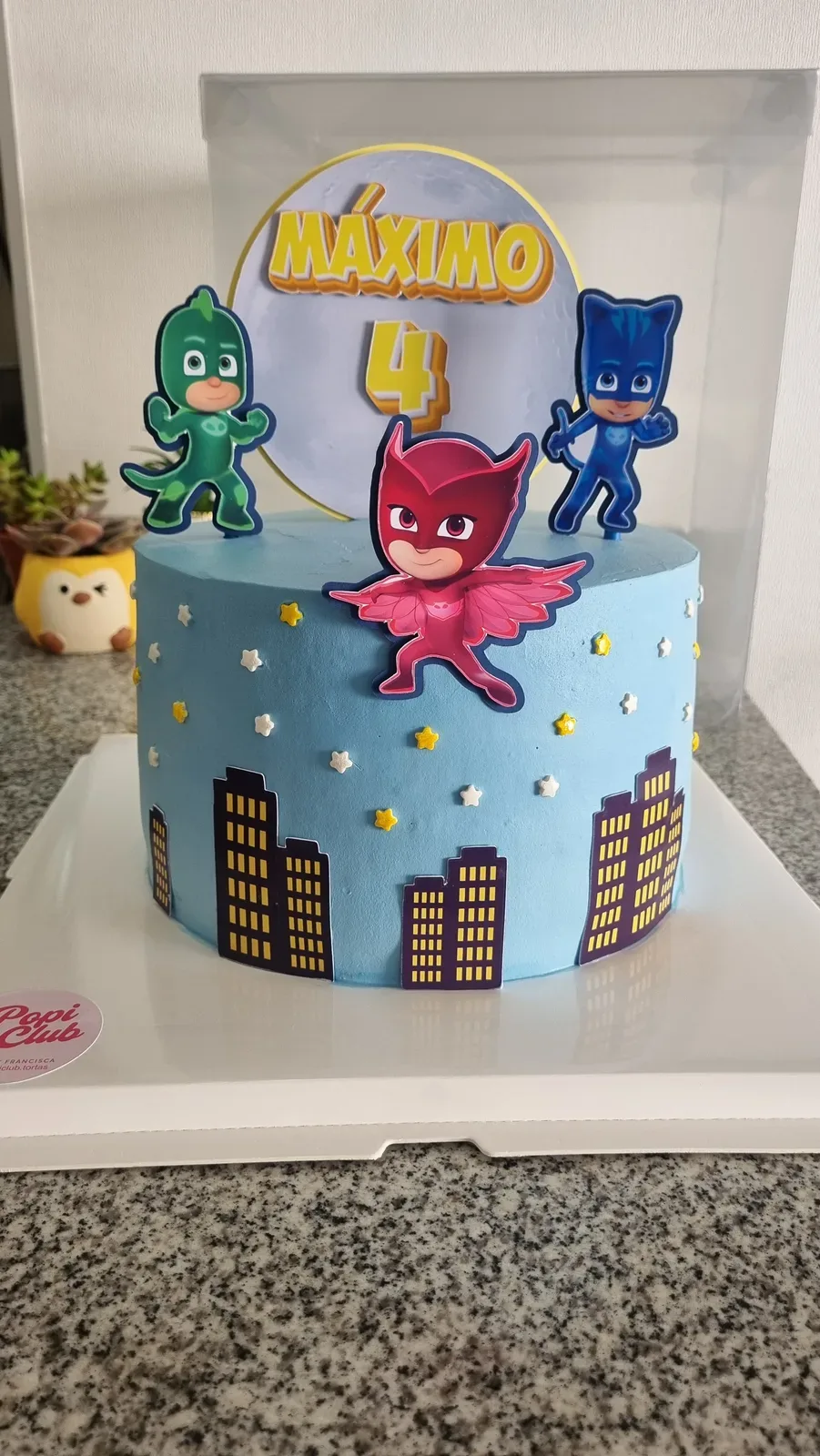 Torta celeste con Héroes en Pijama, nombre Máximo, número 4 y edificios decorativos.