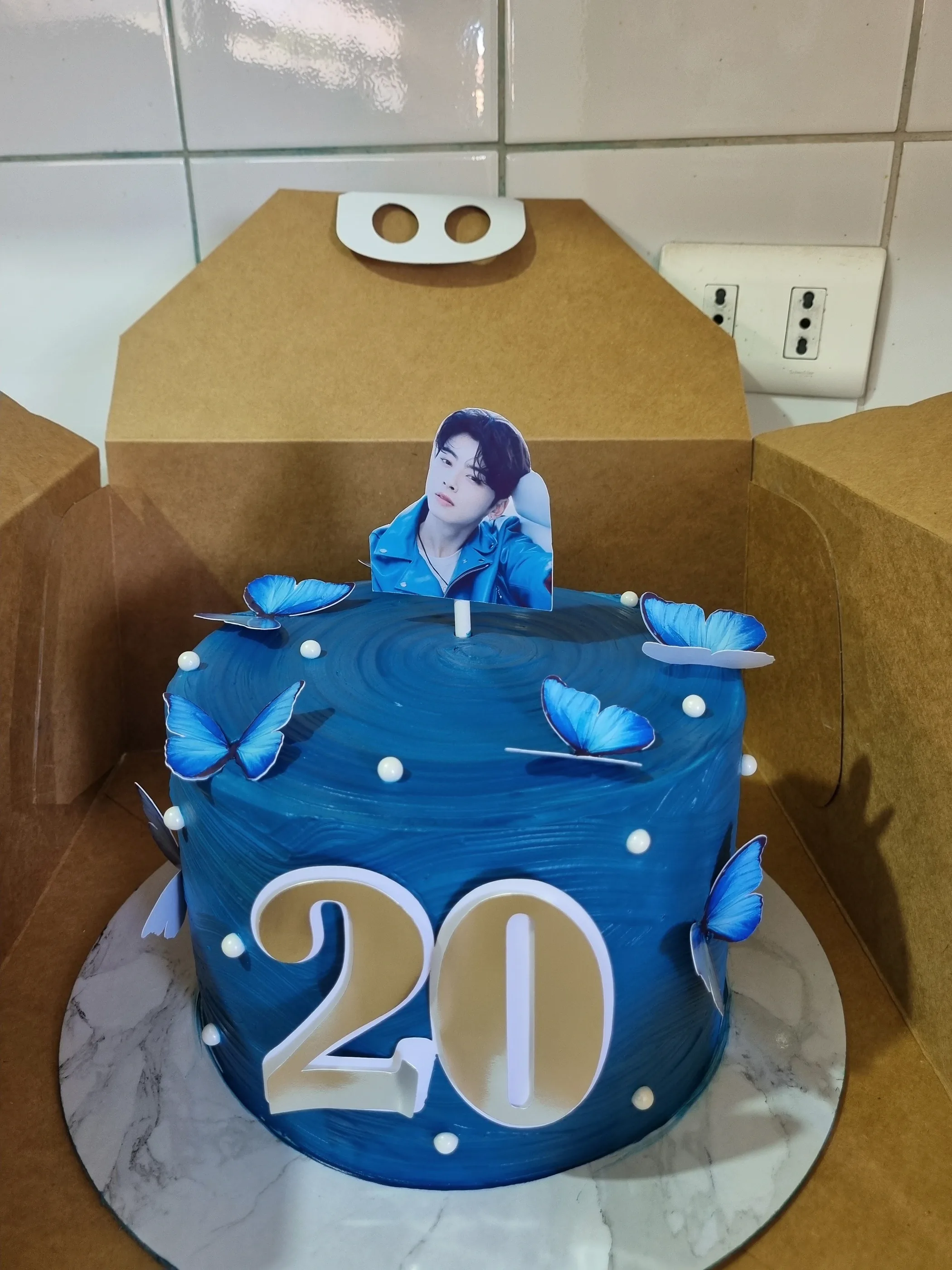 Torta azul con mariposas, perlas, número 20 y topper fotográfico de un cantante sobre base redonda marmolada.