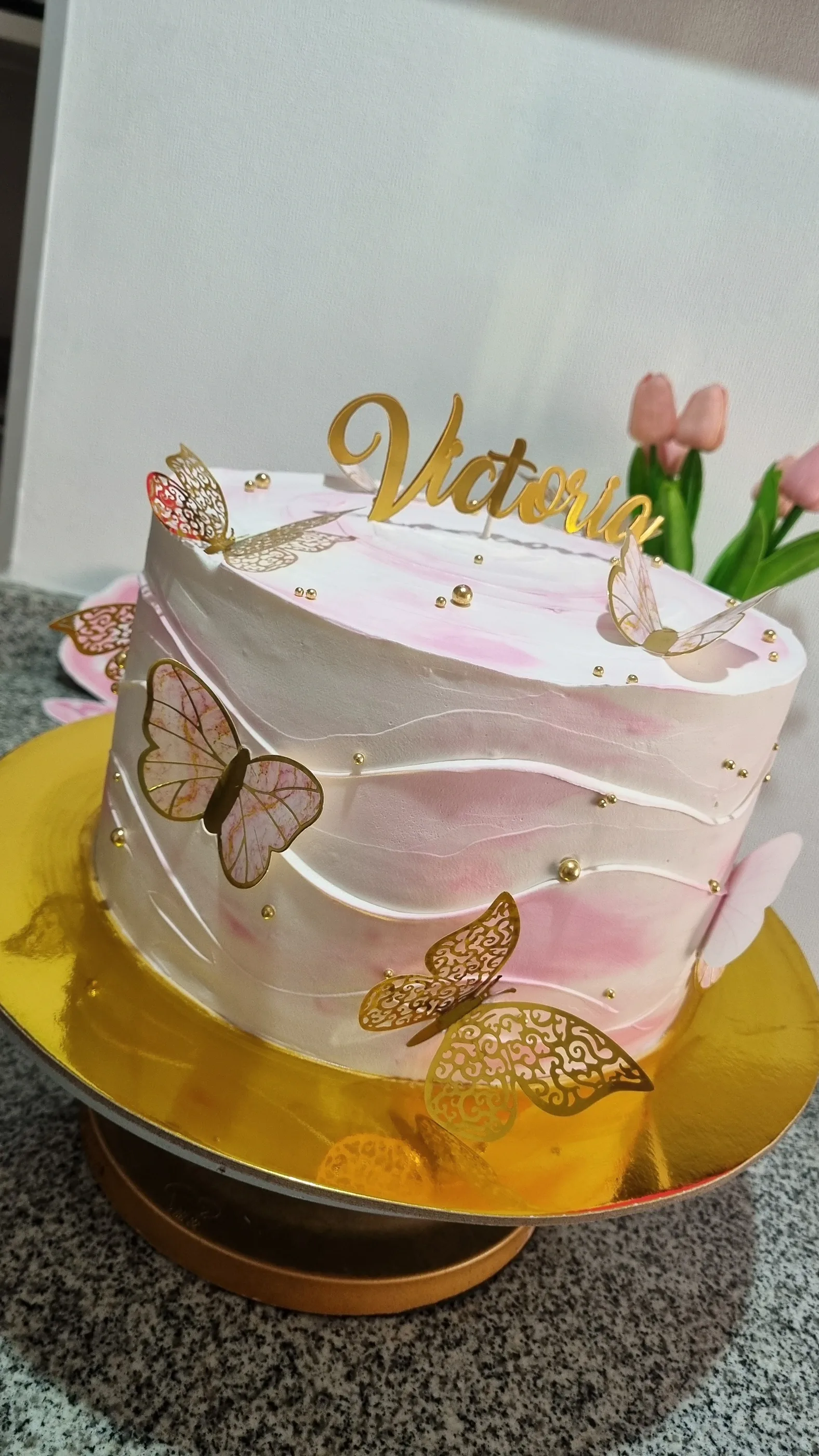 Torta rosa suave con ondas, mariposas doradas y nombre Victoria sobre base dorada.