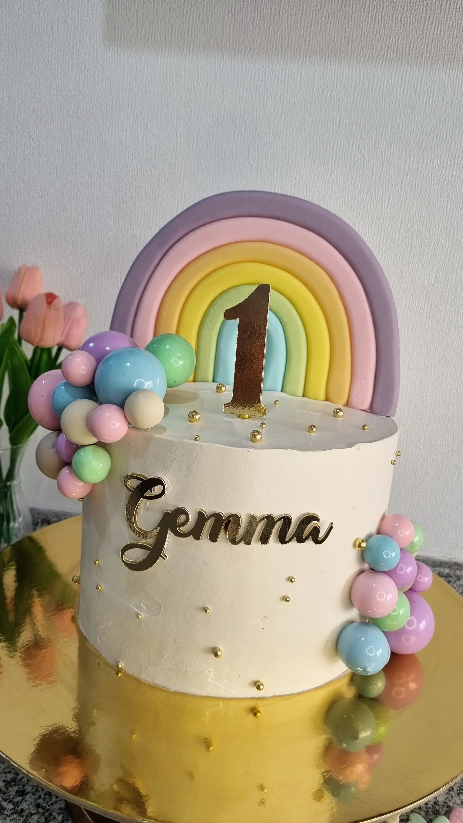 Torta blanca con arcoíris pastel, esferas de colores, nombre Gemma y número 1.
