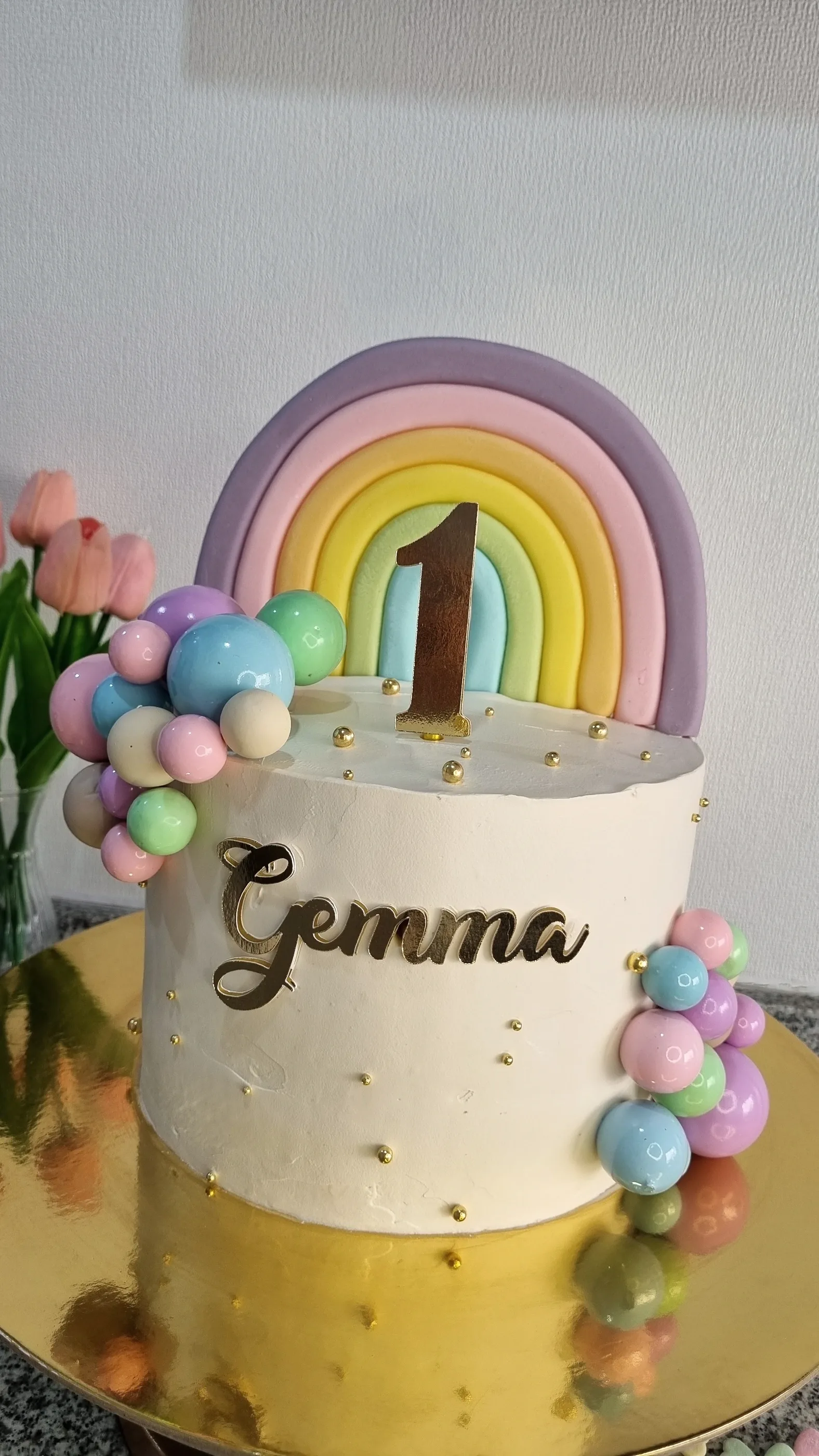 Torta blanca con arcoíris pastel, esferas de colores, nombre Gemma y número 1.