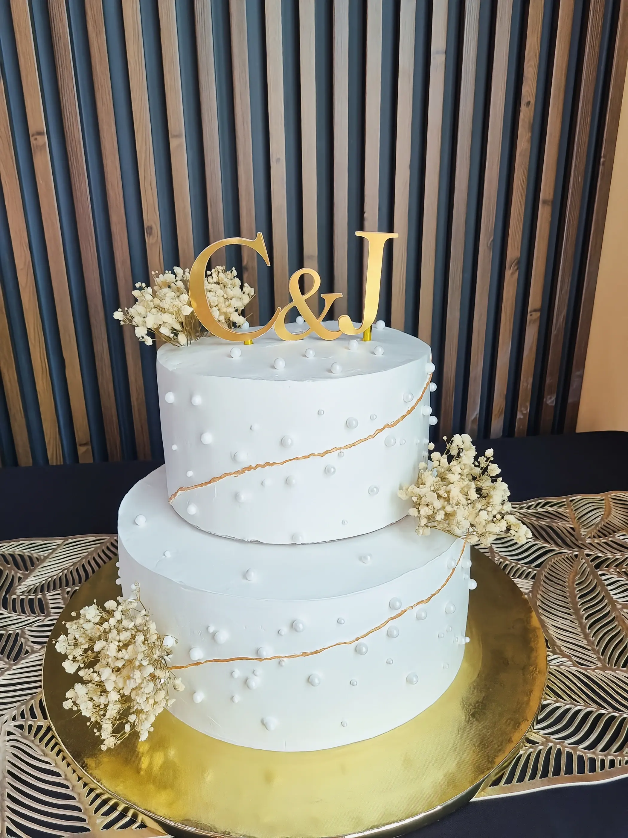 Torta blanca de dos pisos con iniciales C y J doradas, detalles dorados y pequeñas flores claras.