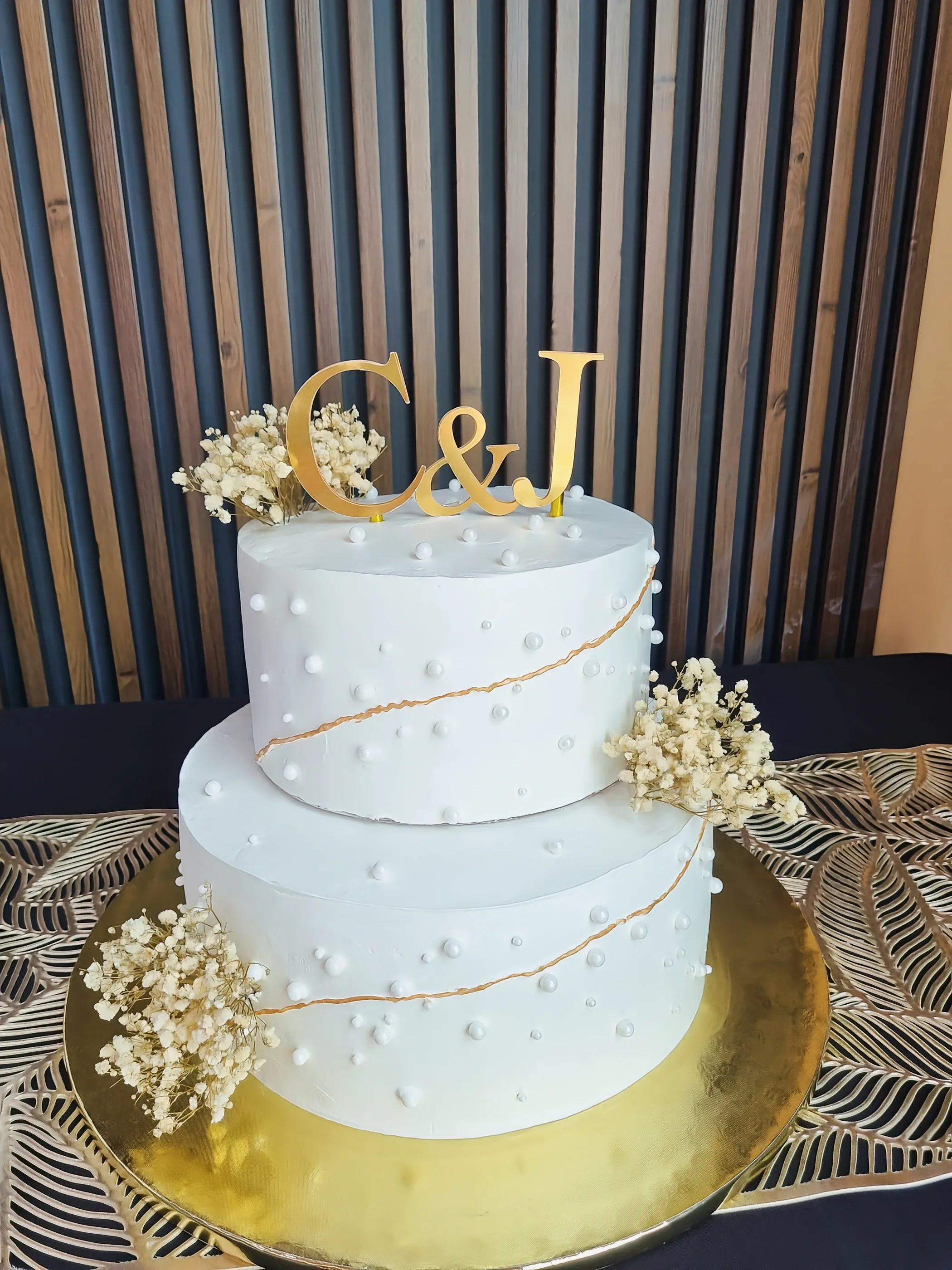 Torta blanca de dos pisos con iniciales C y J doradas, detalles dorados y pequeñas flores claras.